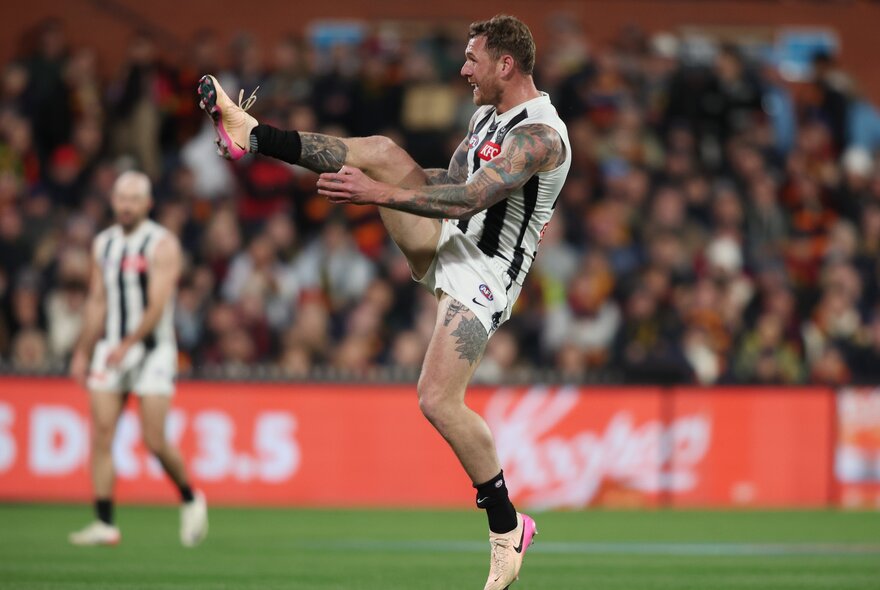 A Collingwood AFL player in mid-air with his leg stretched high after having kicked a football, out of focus fans in the stadium behind him.