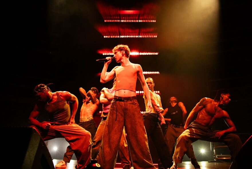 Pop sensation, Troye Sivan, on stage with male back up dancers in a midriff top and baggy jeans, holding a microphone.