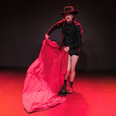 A performer in a black outfit, shorts, and a wide-brimmed black hat stands on a stage with a red floor, holding a large, flowing red cloth like a matador.