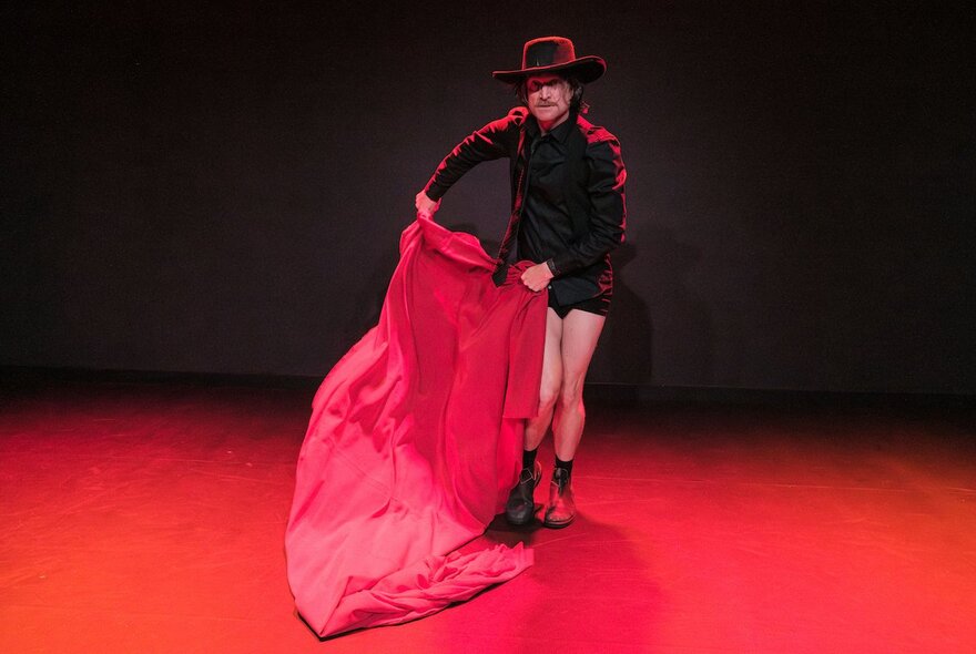 A performer in a black outfit, shorts, and a wide-brimmed black hat stands on a stage with a red floor, holding a large, flowing red cloth like a matador.