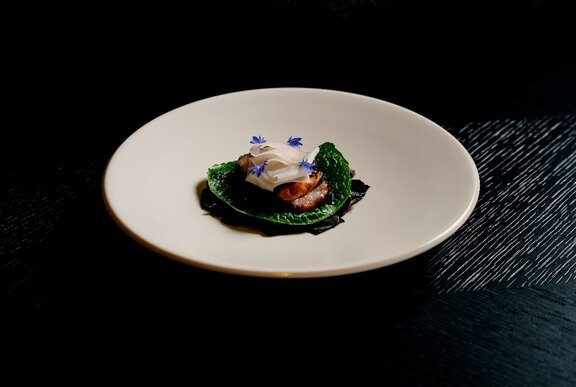 A white bowl with a small Japanese delicacy in the centre, featuring grilled fish with bonito flakes on a green lettuce leaf.