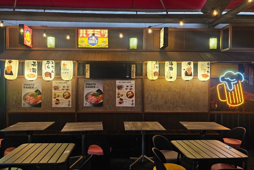 Bar tables in rows next to wall decorated with posters of food, neon beer sign and illuminated pictures.