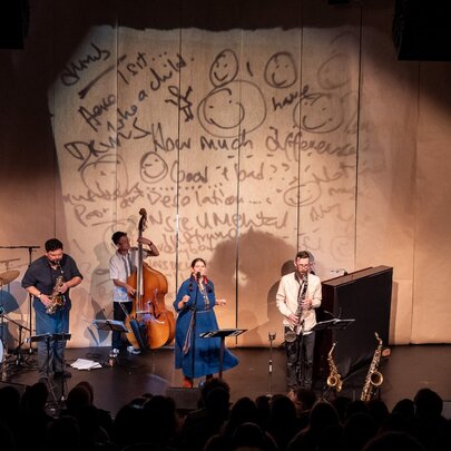 Looking downwards from an audience perspective to a group of musicians on a stage with a projected backdrop of handwritten text. 