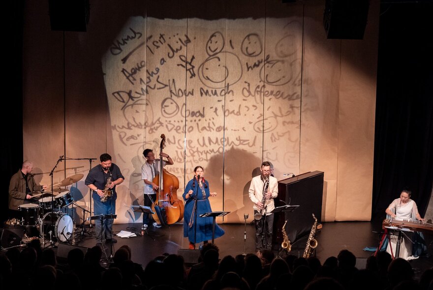 Looking downwards from an audience perspective to a group of musicians on a stage with a projected backdrop of handwritten text. 