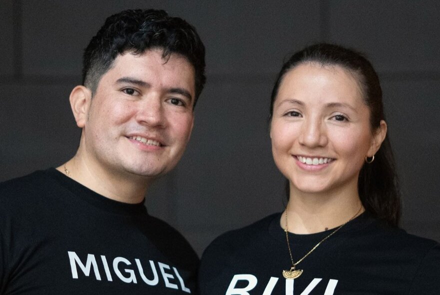 A smiling man and woman stand side-by-side against a dark background, wearing matching black t-shirts with white block lettering.