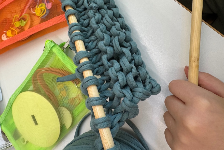 Someone using a large wooden knitting needle to work rows of dark blue-grey T-shirt yarn, working on a white desk with other craft materials on it.