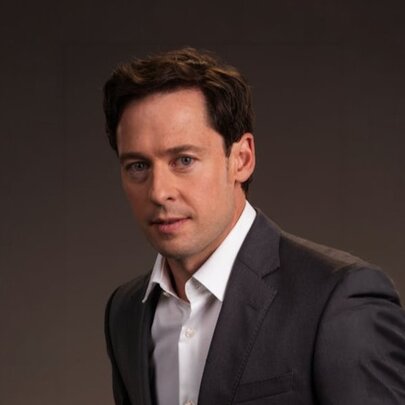 Investigative journalist Nick McKenzie, a man with short dark hair wearing a dark suit jacket and white shirt, posed against a dark brown background.