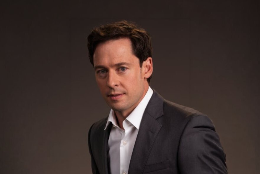 Investigative journalist Nick McKenzie, a man with short dark hair wearing a dark suit jacket and white shirt, posed against a dark brown background.