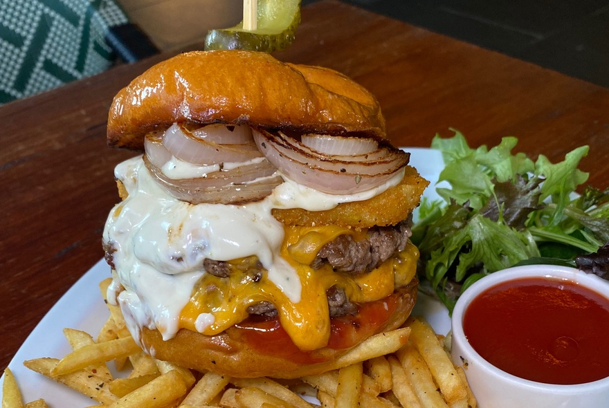 A fully loaded burger with two beef patties, American style cheese, oozing mayonnaise and grilled onions sitting on a bed of fries.