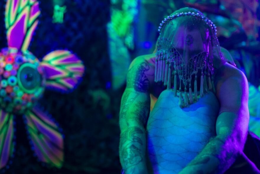 A psychedelic-coloured person in a mermaid costume with a tassled veil over their face, a large psychedelic flower in the background.