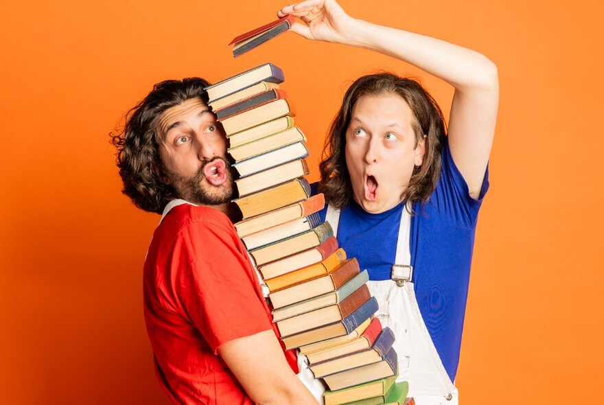 A comedian in white overalls with a bemused and silly expression on his faces, balancing a very large stacked pile of books in his arms while another person keep adding books to the top of the pile.