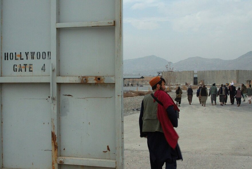 Film still of a man standing outdoors next to a large white structure or partition that has the words HOLLYWOOD GATE 4 written on it, with a row of men and a mountain range in the far distance.