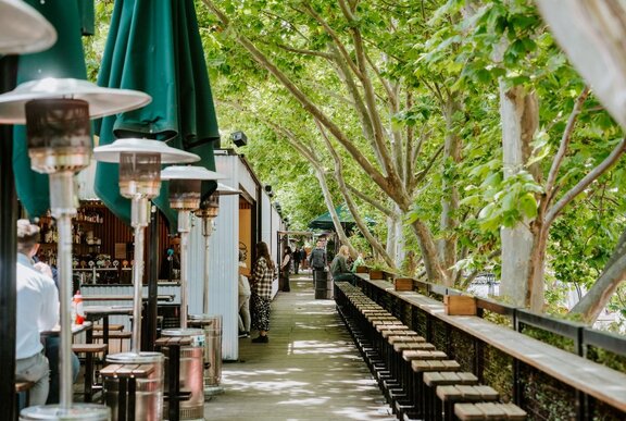 The alfresco terrace of Arbory Bar and Eatery, with a line of stools in front of a long bench table under leafy green trees.