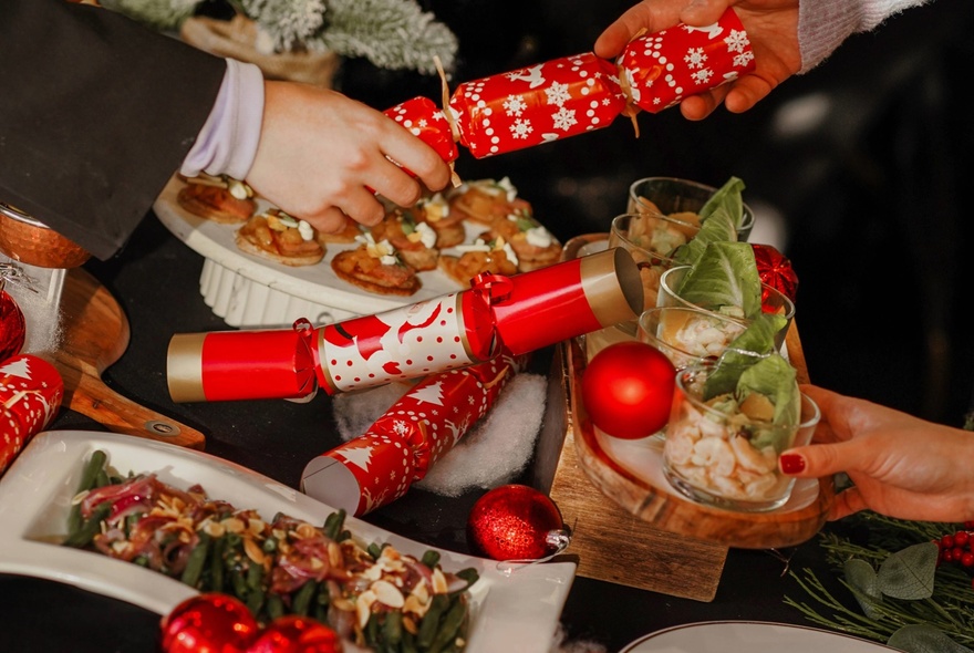 A festive high tea buffet spread on a table, with people passing Christmas bon bon crackers and plates of food to each other.