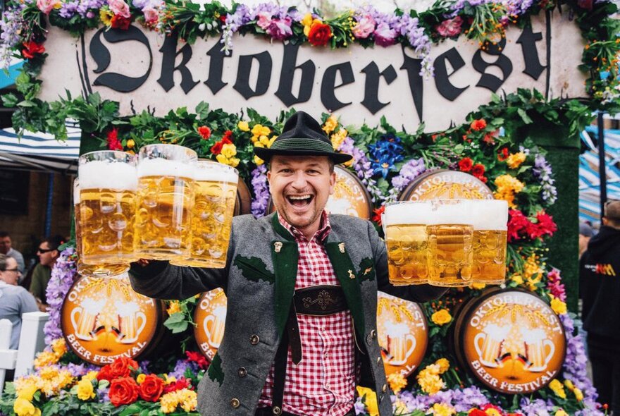 A very happy man in a German-themed outfit holding up 6 beer steins in front of an Oktoberfest sign. 