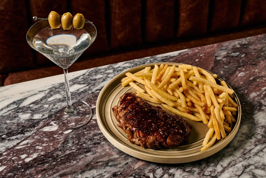 A plate of steak frites and a martini with three olives on a marble tabletop. 