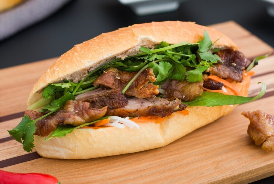 A large bahn mi with pork, coriander and carrot, on a wooden board.