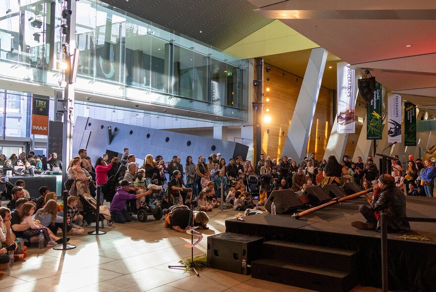 The foyer of Melbourne Museum, with people seated on the floor watching and listening to a performance on a stage.