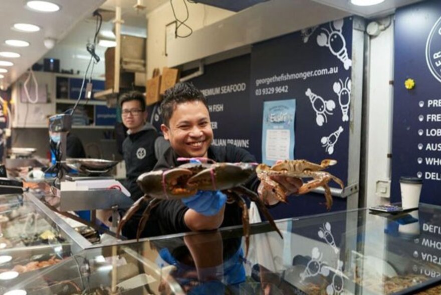 A smiling fishmonger at Queen Victoria Market holding two crabs in his hands over the glass counter of the stall, towards an out of sight customer.