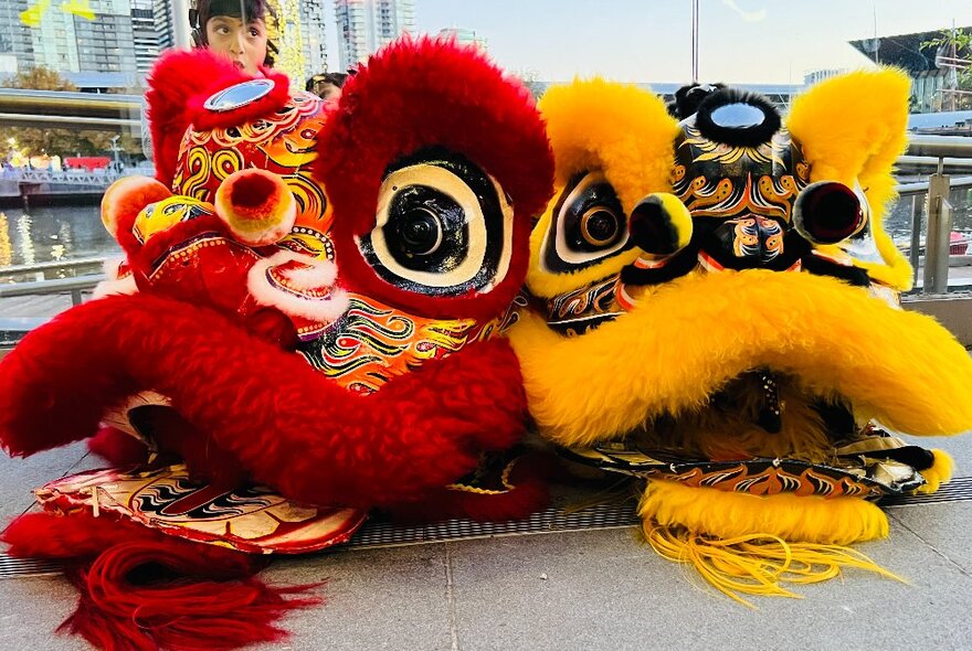 Red and yellow lion dance figures on a walkway overlooking the water and towers of Docklands.