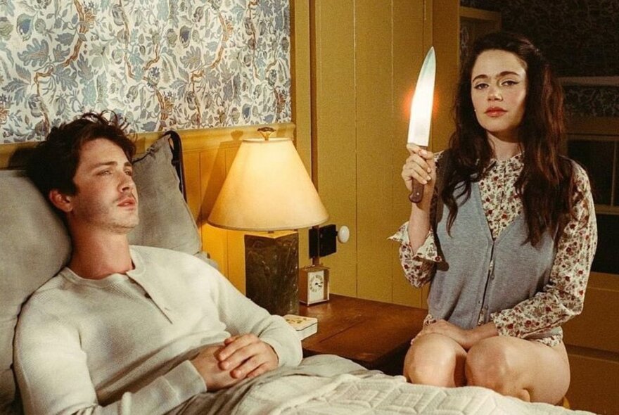 A still from the film, Oh, Hi! showing a young woman holding a knife and sitting next to a bed with a young man lying in it.