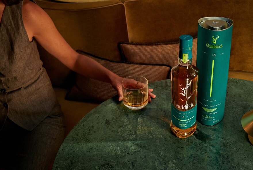 Hand holding a glass of whisky next to a Glenfiddich whisky bottle and container.