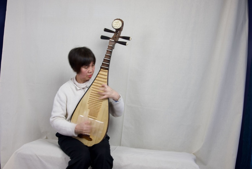 A seated person playing a traditional Chinese pipa, a four-stringed plucked instrument with a pear-shaped wooden body, against a white background.