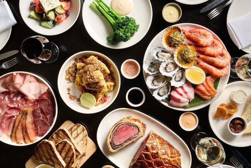 Looking down on a table with many plates, large and small, of beef wellington, veggies seafood and condiments.