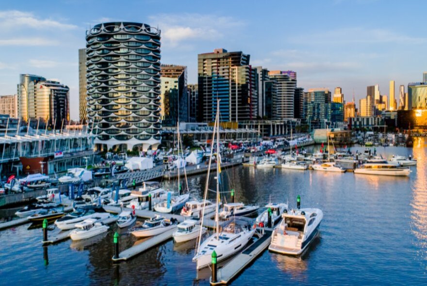 Docklands marina with boats moored by the walkway and city towers in the background.