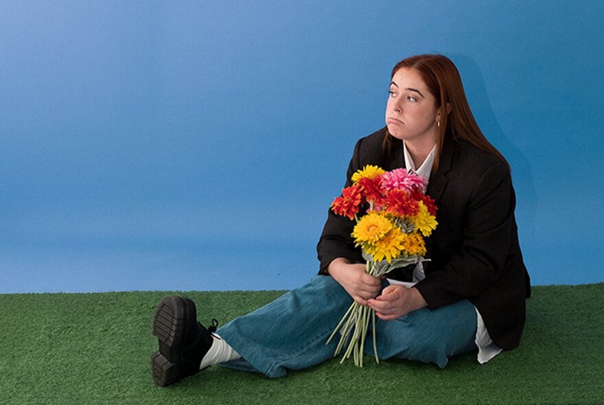Woman seated on fake grass holding a bunch of flowers, looking grim.