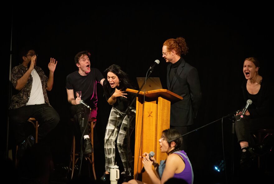 An ensemble cast of six actors on a stage, one person standing at a lectern in front of a microphone.