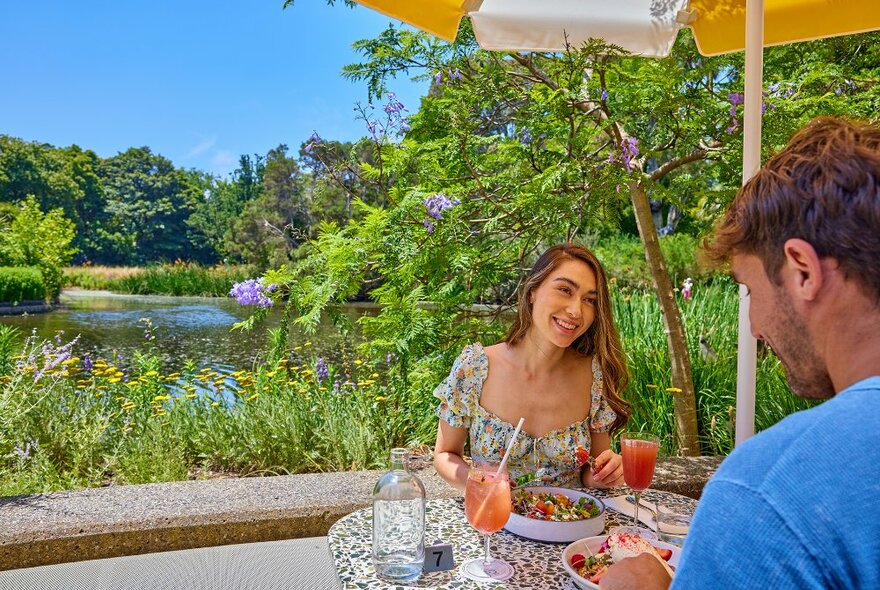 A couple sharing a meal and cocktails by a garden lake.