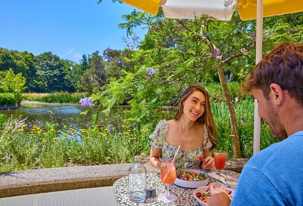 A couple sharing a meal and cocktails by a garden lake.