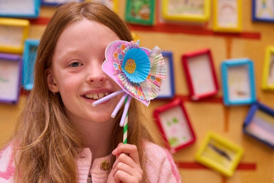 A girl holding a handcrafted paper flower in front of her face, smiling, tens of coloured picture frames on the yellow wall behind her.
