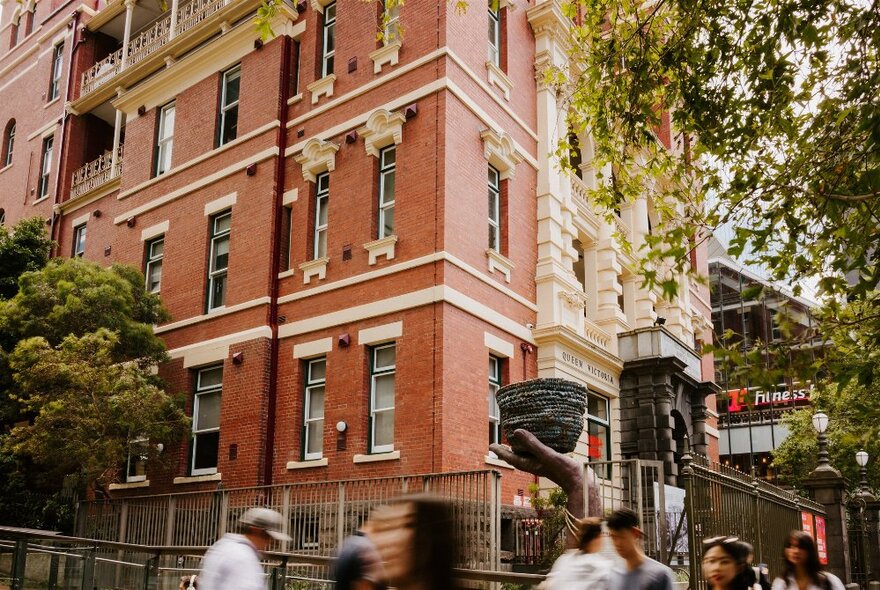 Corner view of the red and cream brick exterior of the Queen Victoria Women's Centre with trees  and walking pedestrians in the foreground.