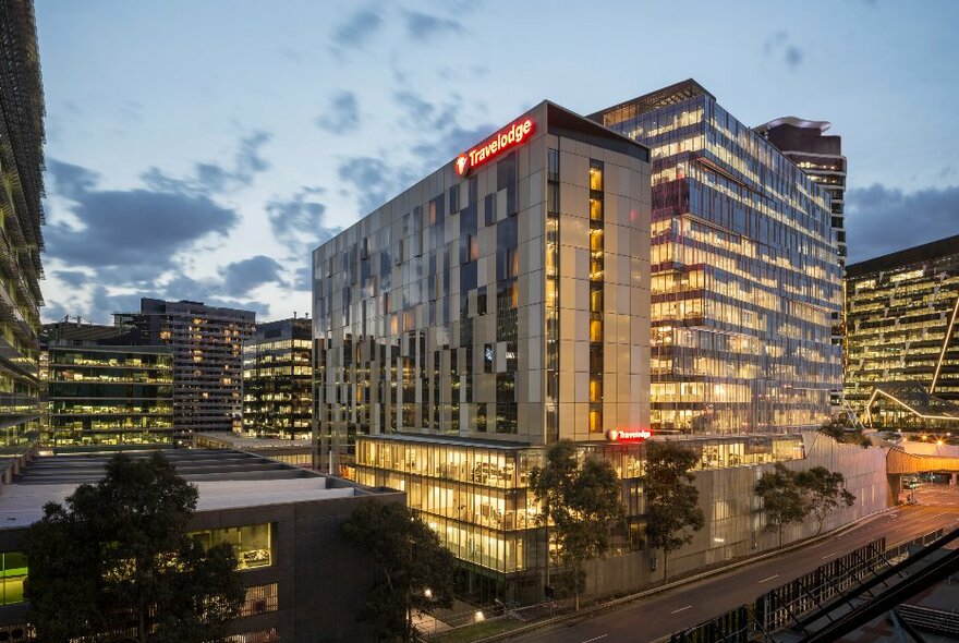 Mid-level hotel with glass illuminated walls, surrounded by similar lit-up buildings at dusk.