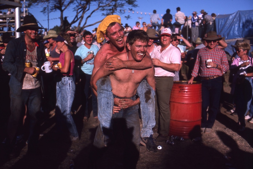 A photograph of a bush party in outback Queensland, where one shirtless man is carrying another on his back, while onlookers laugh.