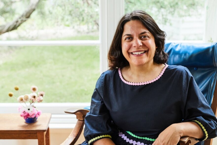 Author Jaclyn Crupi smiling and seated in an armchair by a window, a coffee table next to her with a small floral arrangement on it.