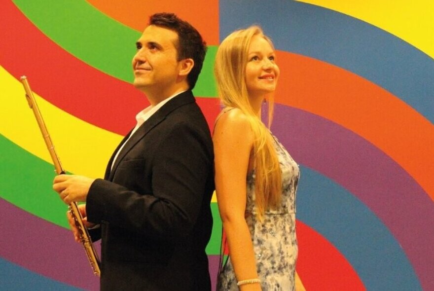 Male musician holding a flute standing back to back to a blonde woman wearing a blue and white sleeveless dress against a multicoloured striped retro background.