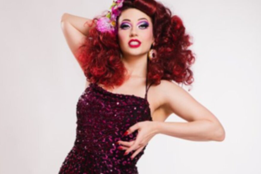 A burlesque performer striking a pose wearing a sequined gown, her red hair arranged in a voluminous hair-style around her face.