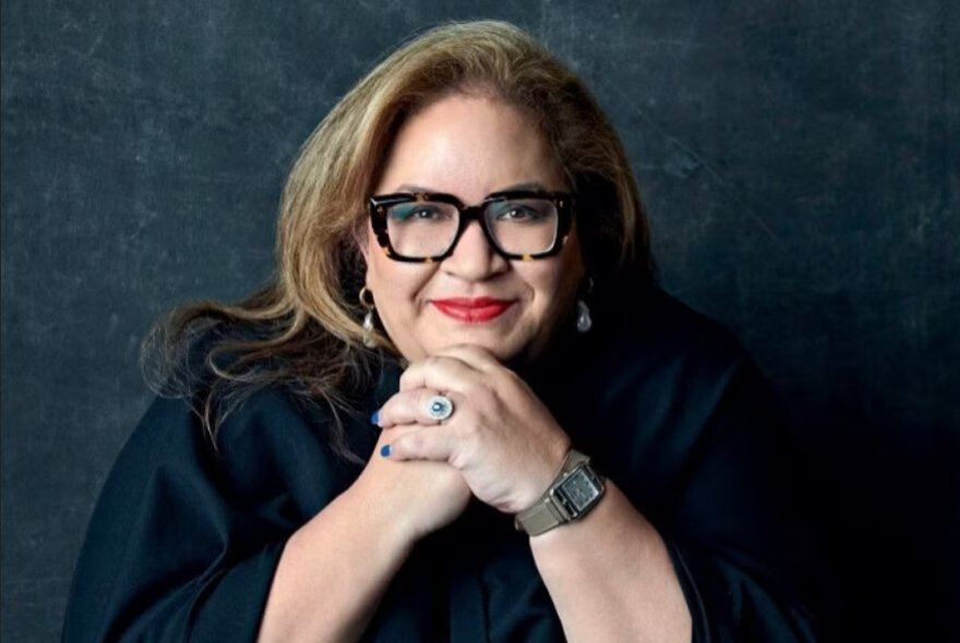 Woman with glasses, her hands folded under her chin.