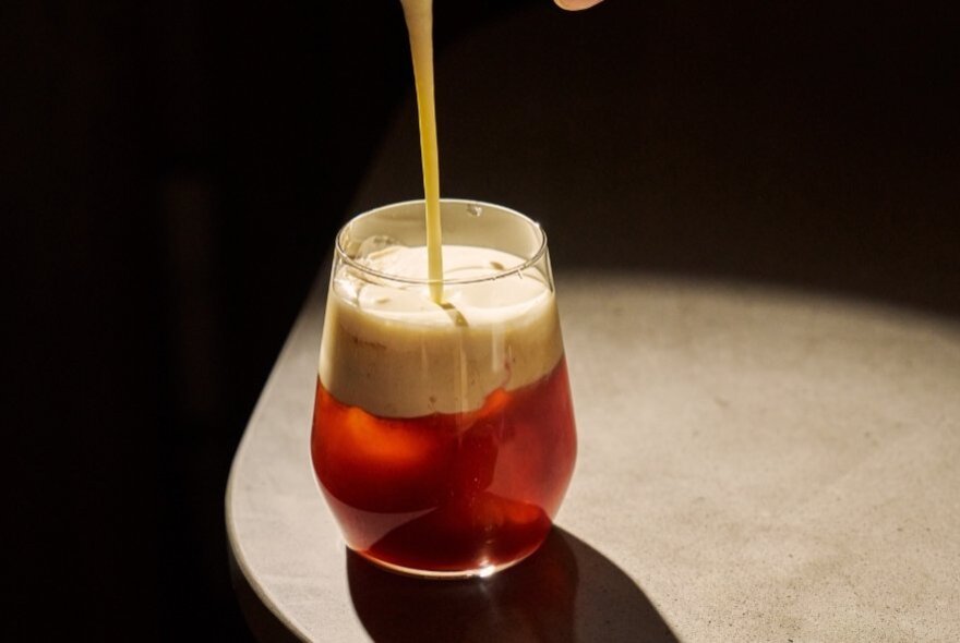 A frothy drink being poured into a glass tumbler that rests on a dark surface.
