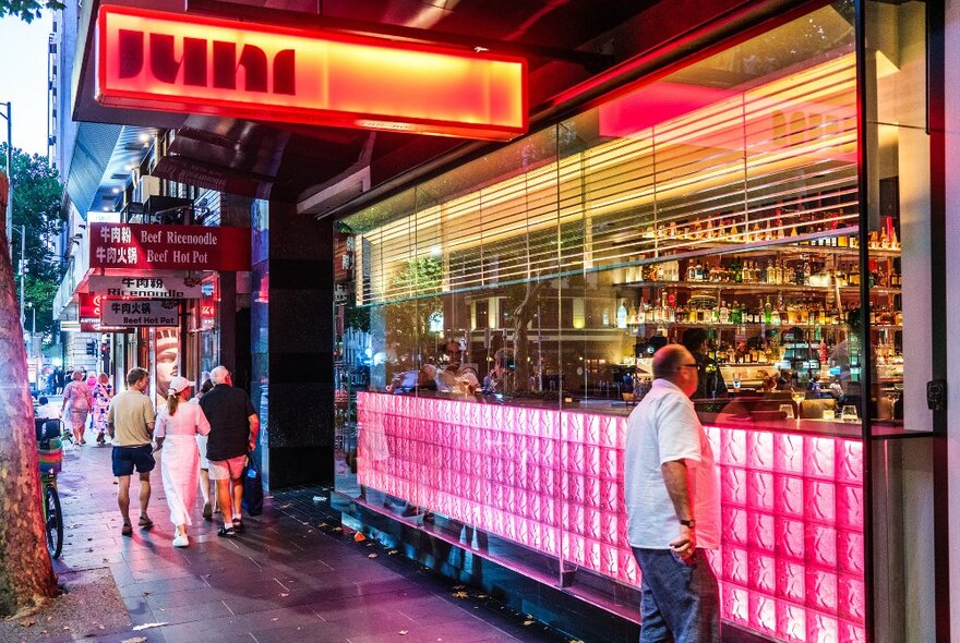 A man is looking into a window of a neon-lit restaurant. There is a sign saying JUNI. 