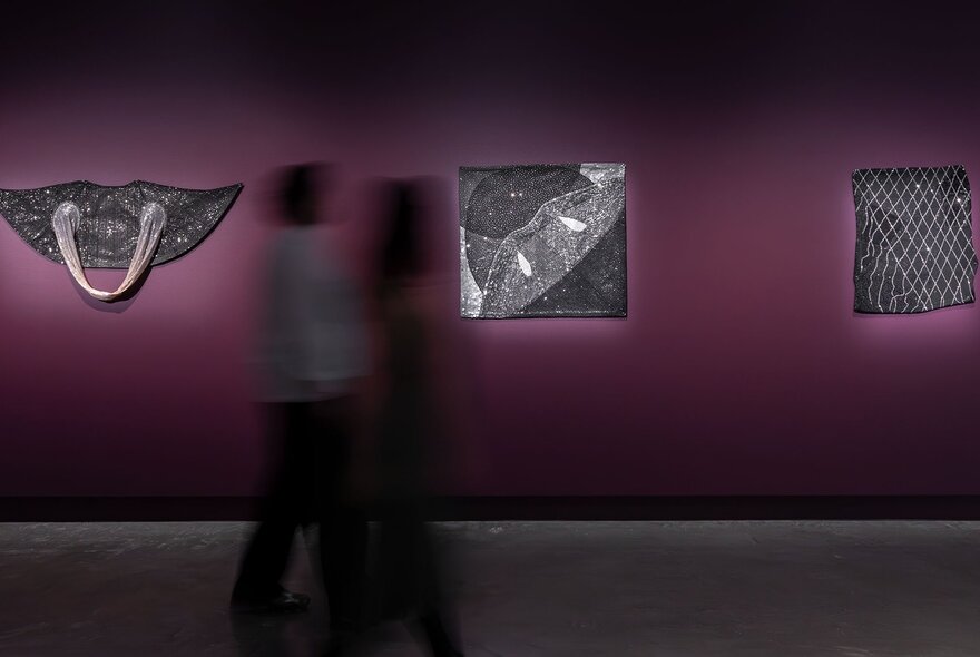 People walking past grey artworks displayed on purple walls in a gallery.