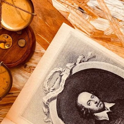 An antique-style wooden balance scale beside an open book featuring a portrait engraving of William Shakespeare.