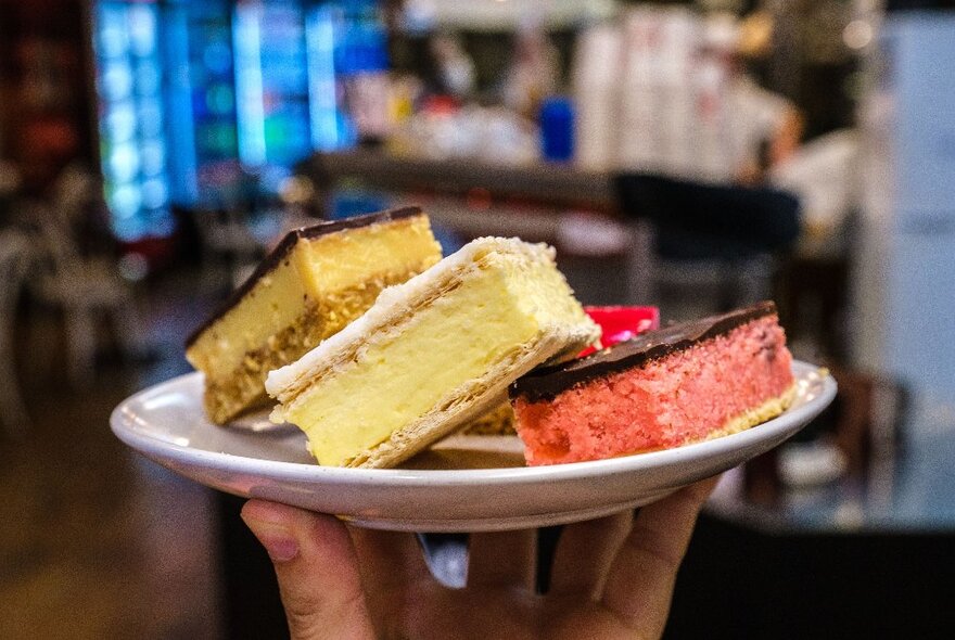 A hand holding up a plate of dessert slices including a vanilla slice, a raspberry slice and a caramel slice.