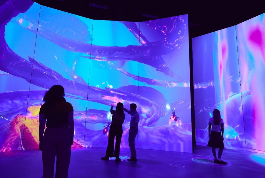 Silhouettes of people looking at giant projections in a room with screens, the images are swirly and blue and purple tones. 