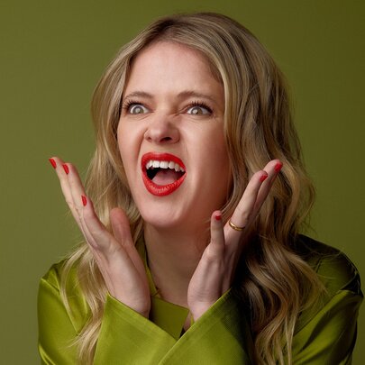 A young person with long blonde hair, wearing a  green satin top and making an animated, expressive gesture with hands up near their face against a solid olive-green background.