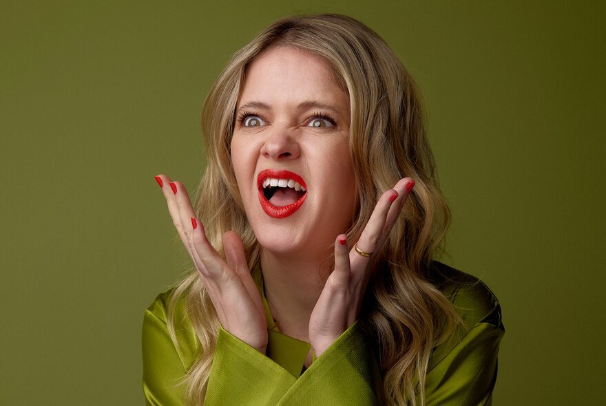 A young person with long blonde hair, wearing a  green satin top and making an animated, expressive gesture with hands up near their face against a solid olive-green background.