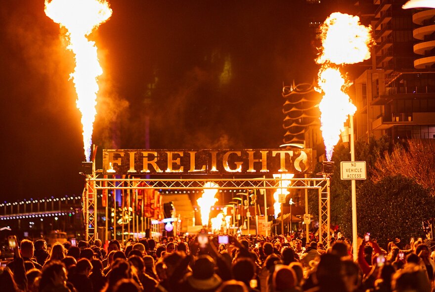 A crowd of people are at a winter festival they are walking under a large sign saying firelight which has flames shooting out from either side
